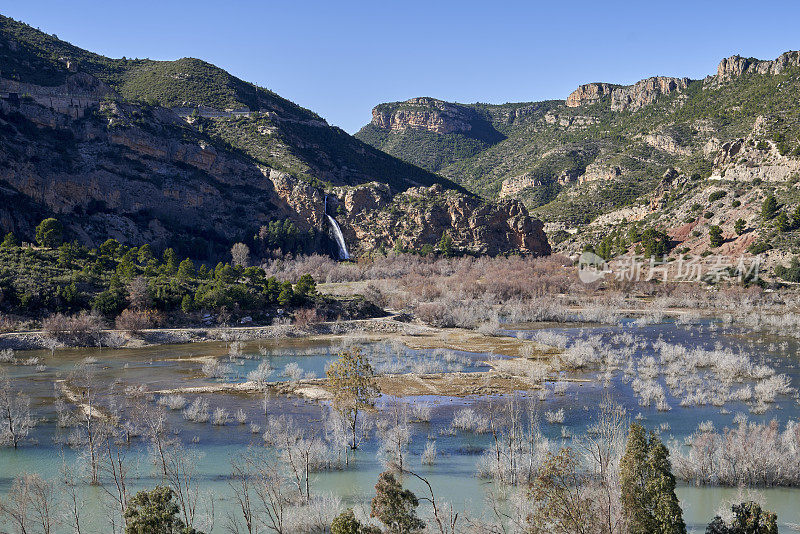 Beautiful landscape of the waterfall near the old town of Domeño on a rocky mountain and the Turia river with trees in the foreground, in Valencia, Spain